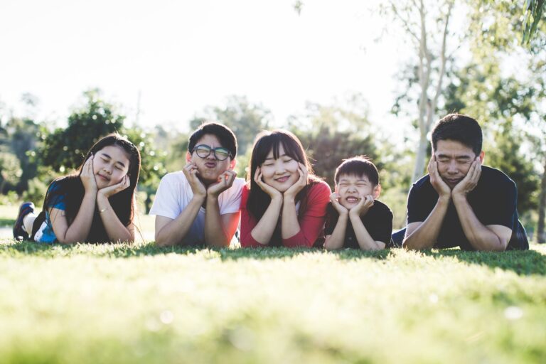 A family of five lies in their bellies on the grass, faces rested in their palms, and makes silly faces at the camera.