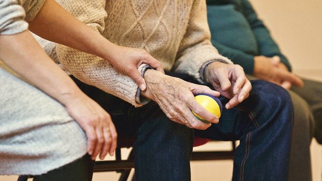 A woman grips the hand of an older man who holds a squeeze ball. We only see them from the waist down.