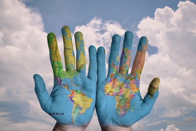 A pair of hands are painted to look exactly like a map of the globe and they are held up against a blue sky with white puffy clouds.