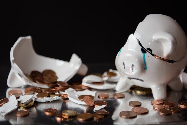 A white piggy bank sits broken in half, a tear running down his little piggy face, as all his coins lie strewn about around his broken ceramic parts.