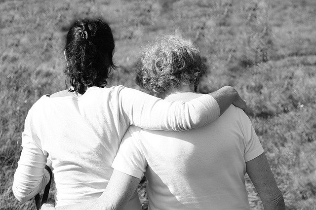 Two women are turned away from us, walking with arms around each other, in a field.