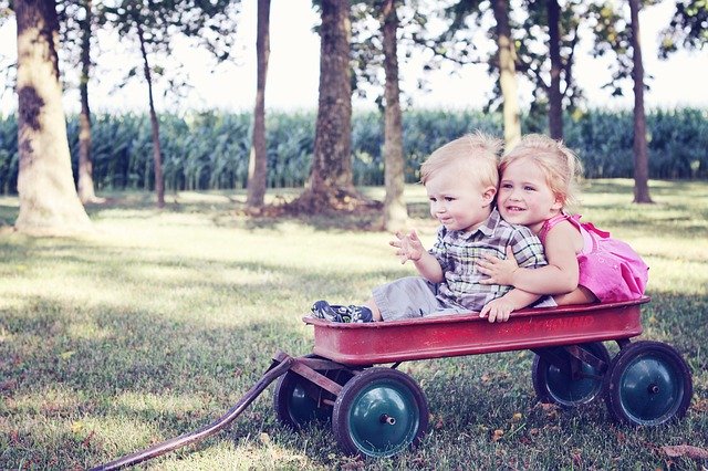 A young boy and girl sit close together in a red wagon in the park. She is hugging him.