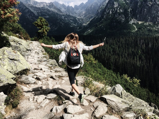 A young woman wearing a backpack skips down the side of a mountain, her arms flung out to her sides.