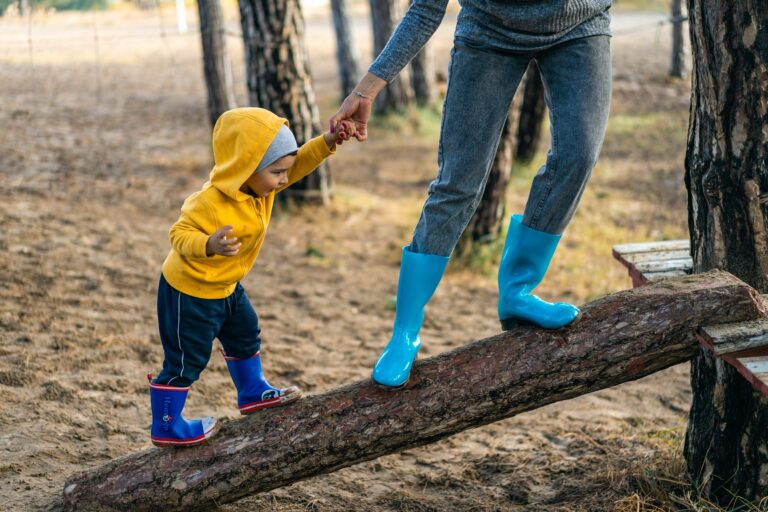 A woman in bright blue rain boots carefully helps a toddler, also in blue rainboots, walk across a fallan log ramp. He wears a bright yellow jacket with the hood over his head.