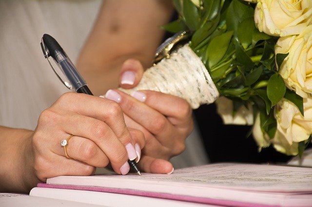 A woman with a wedding ring on, holding a boquete of flowers, signs a document.